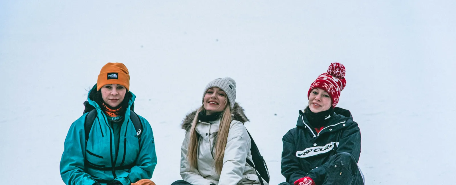 Three people sitting in the snow wearing winter jackets, hats, and gloves smiling at the camera outdoors.