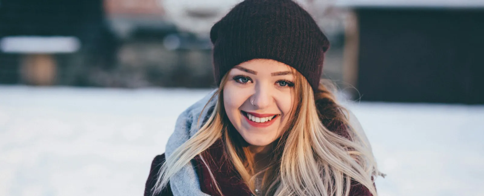 Smiling young woman wearing a maroon beanie, scarf, and coat standing outdoors in snowy winter setting.