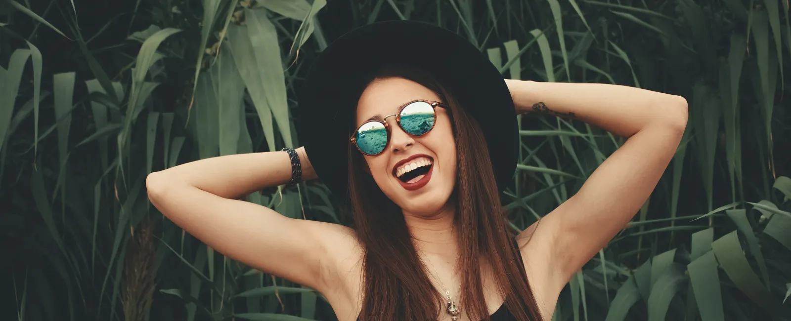 Happy young woman in black dress and hat wearing sunglasses smiling with hands behind head in green foliage.