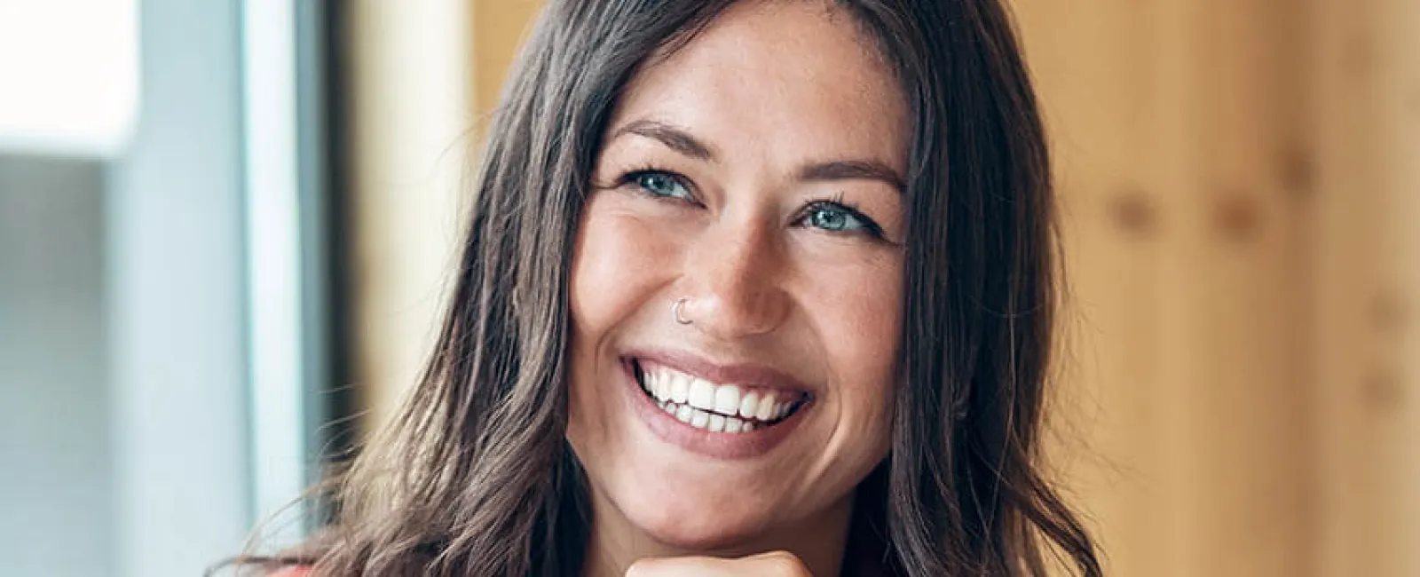 Smiling young woman with blue eyes and brown hair wearing red jacket sitting indoors by window.