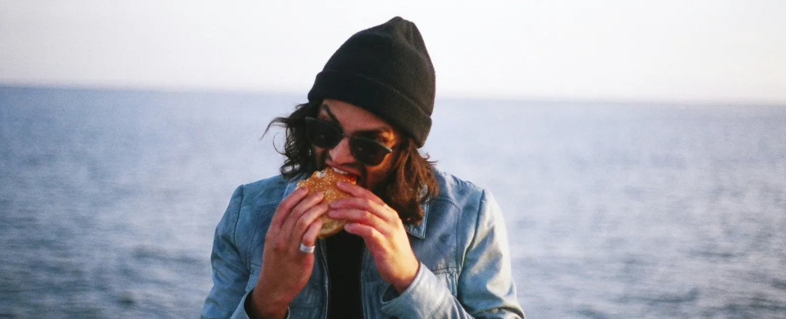 Person wearing sunglasses and a beanie eating a sandwich by the waterfront with railing and ocean in background