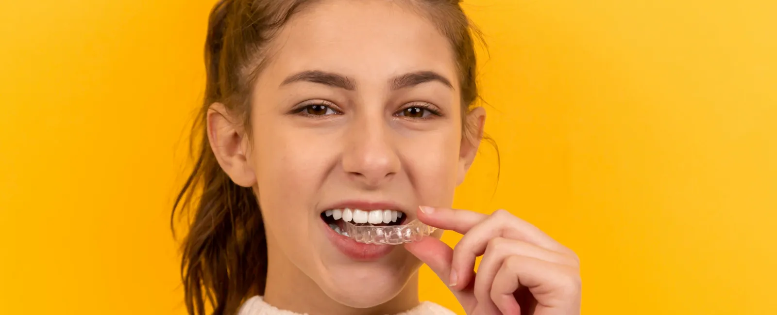 Young woman smiling as she places a clear dental aligner on her teeth against a yellow background