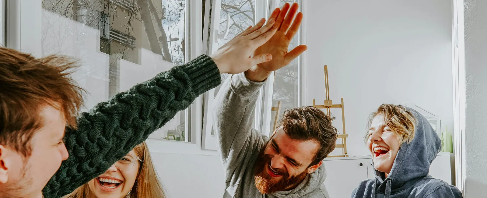 Four friends in cozy clothes enjoying tea and laughing by a window, sharing a joyful high-five indoors.