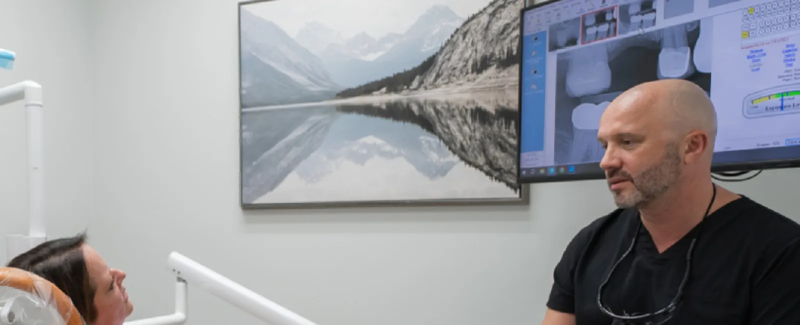 Dentist consulting female patient in dental office with X-ray images on screen and mountain art on wall