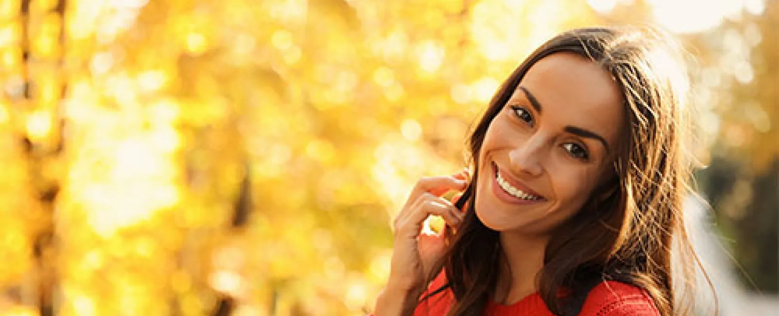 Smiling woman in a red sweater standing outdoors with blurred autumn foliage in the background