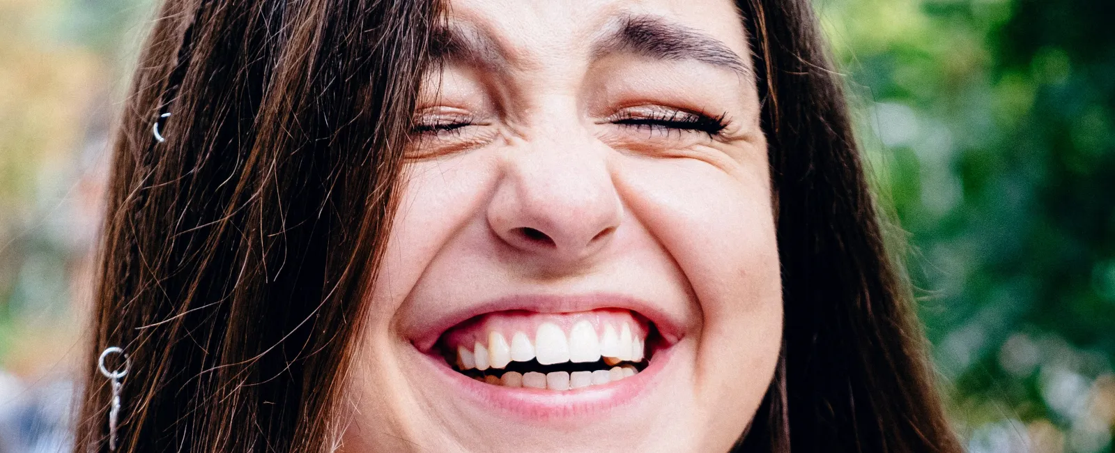 Young woman with long brown hair laughing joyfully outdoors wearing a black tank top with a blurred green background