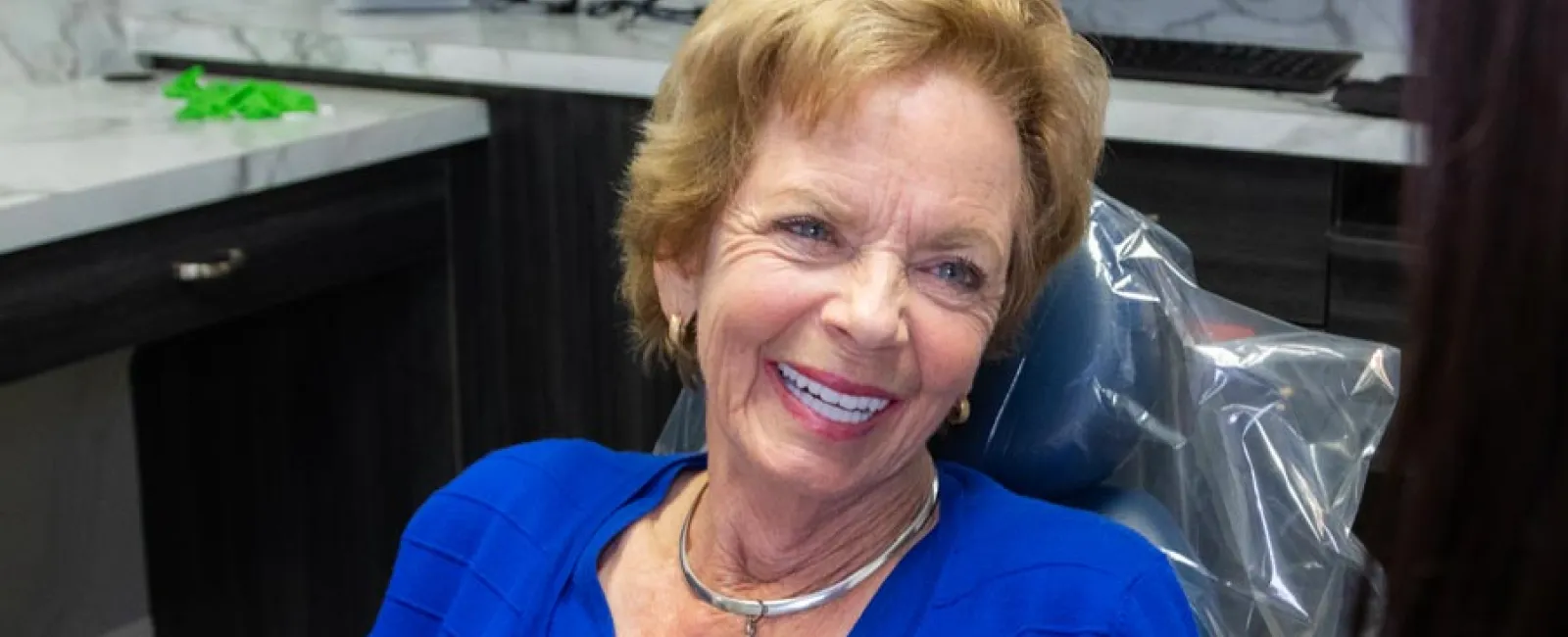 Smiling elderly woman in a blue top sitting in a dental chair during a consultation at the dental office.
