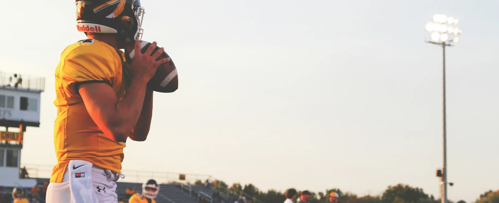 High school football player in yellow jersey prepares to throw ball during practice on field at sunset.