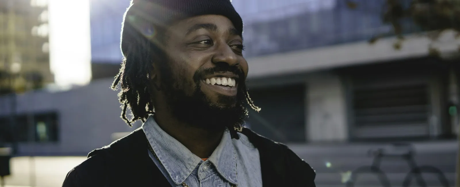 Smiling young man with dreadlocks wearing a beanie and denim shirt outdoors in urban setting with sunlight.