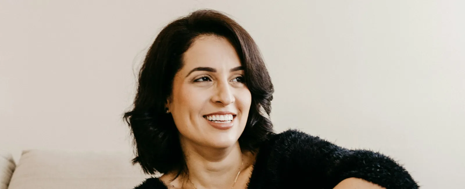 Smiling woman in black sweater and white pants sitting barefoot on striped couch indoors.
