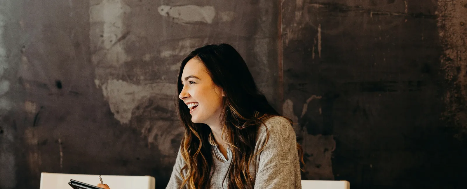 Smiling woman in gray sweater holding a tablet and pen sitting at a wooden table with dark textured wall background