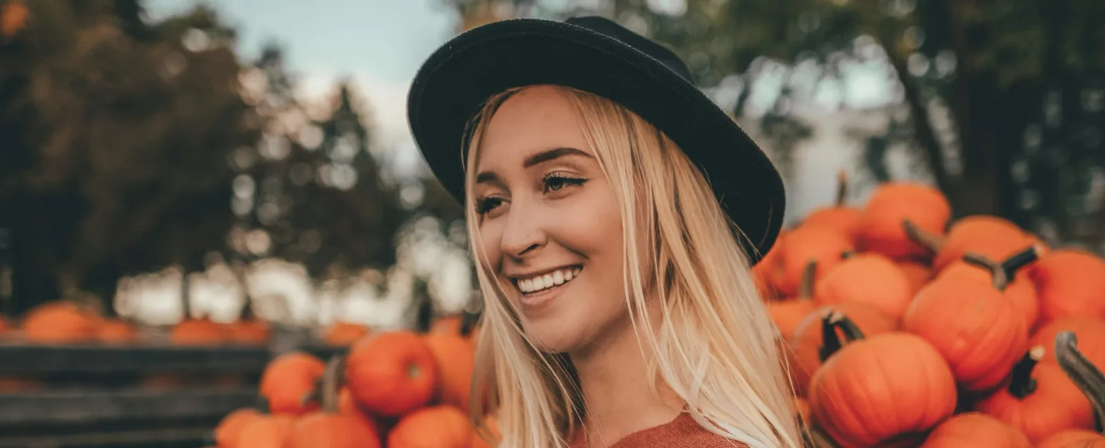 Smiling woman in striped sweater and hat holding a pumpkin by a wooden cart filled with pumpkins in autumn.