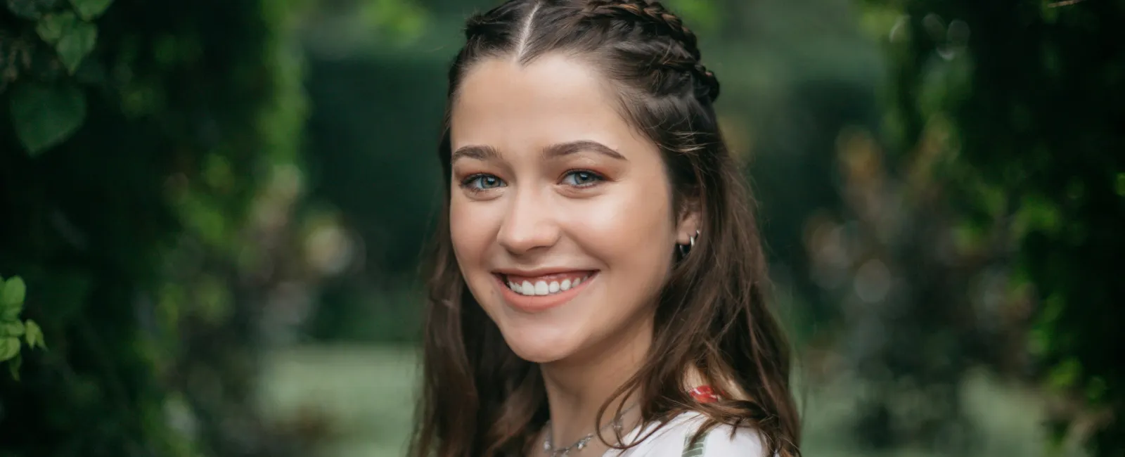 Smiling young woman with braided hair in floral dress posing outdoors with lush green background.