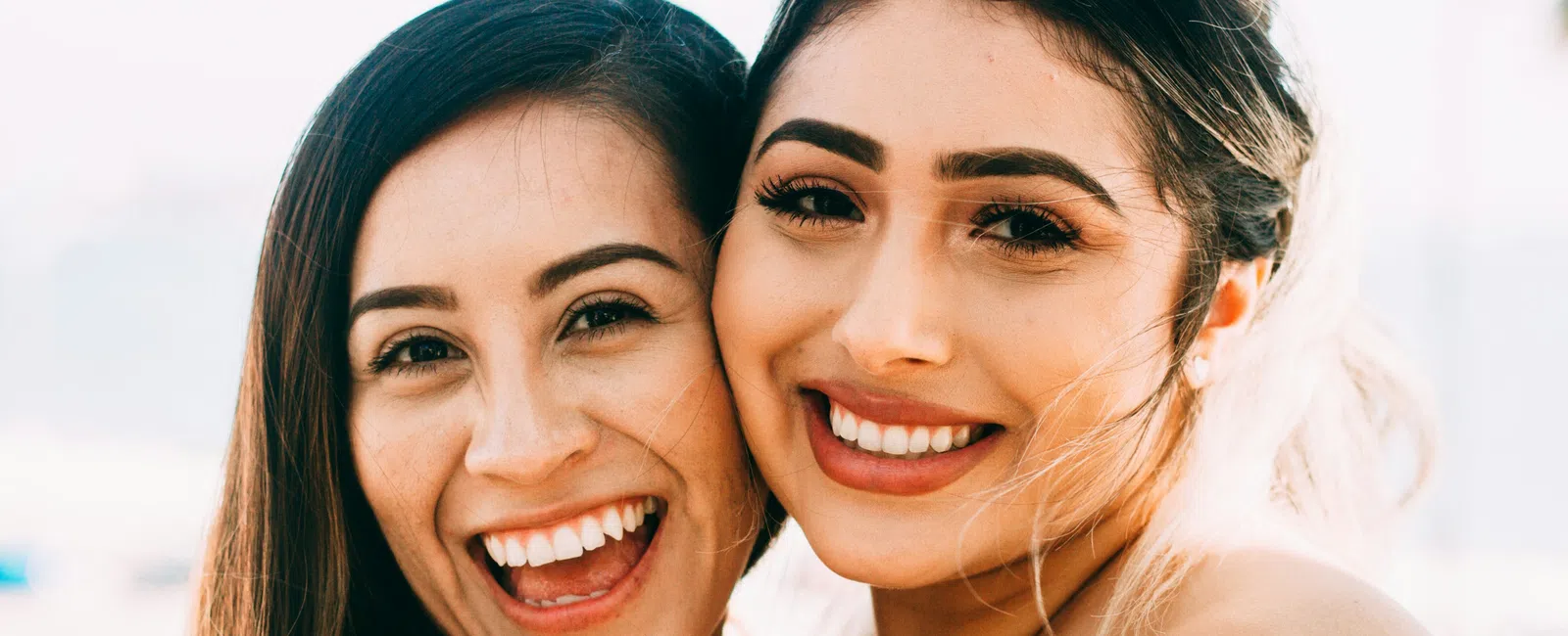 Two young women with bright smiles posing closely together in natural light outdoors.