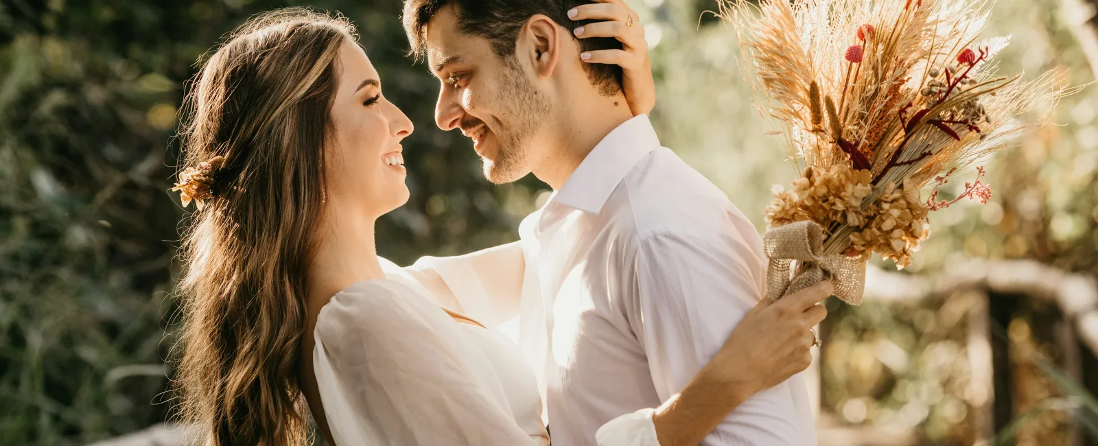 Couple in white clothing embracing outdoors with a dried flower bouquet on a sunny day.