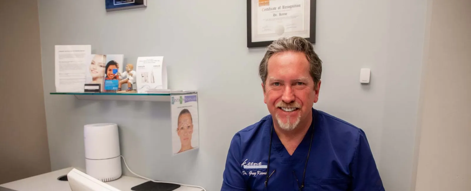 Smiling male doctor in blue scrubs sitting at desk with computer in a medical office setting.
