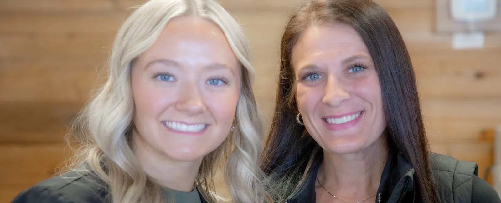 Two smiling women with blonde and brunette hair posing indoors in casual attire against wooden wall background