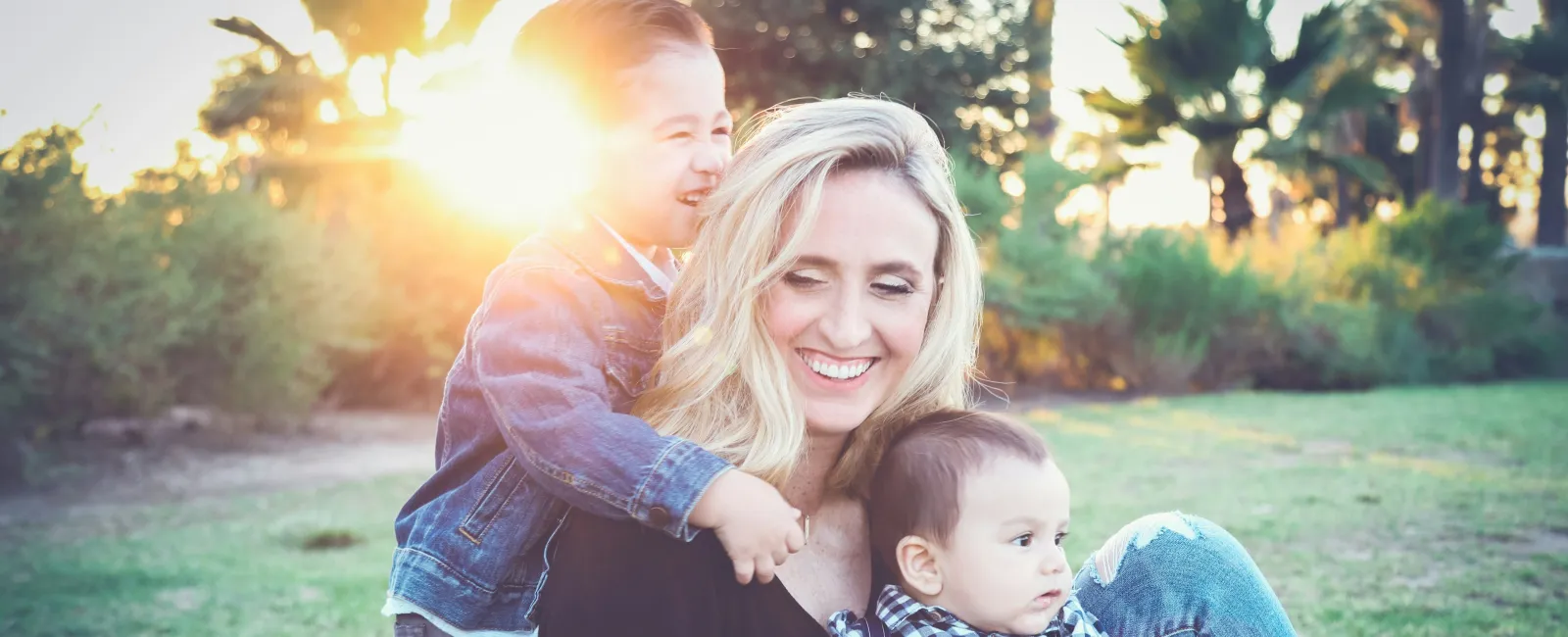 A joyful woman with two children, smiling in a sunny park surrounded by greenery and palm trees.
