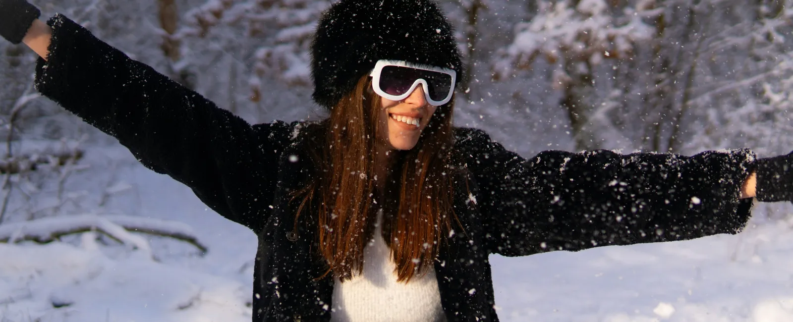 Joyful woman wearing winter clothes and sunglasses enjoying falling snow outdoors in a snowy forest.