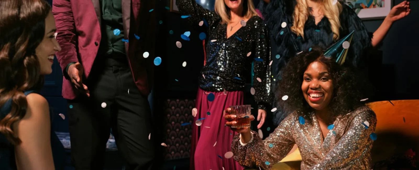 Diverse group of friends celebrating with drinks and confetti at an indoor party with balloons and decorations.