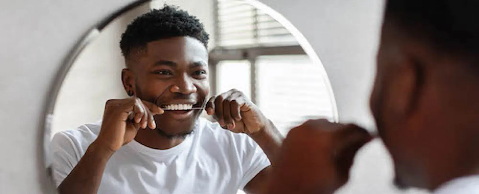 Smiling man flossing teeth in front of a round mirror in a bright bathroom.