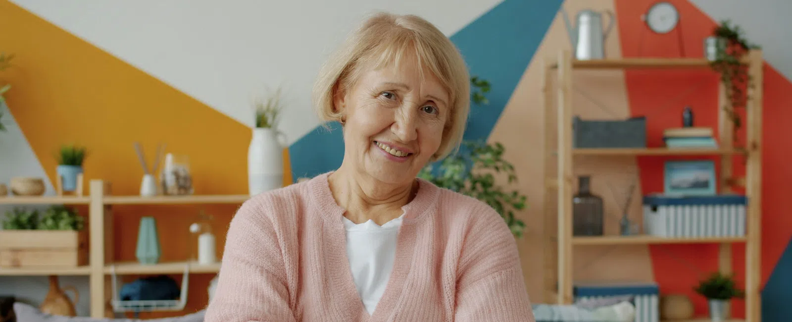 Smiling elderly woman in pink sweater standing with arms crossed in cozy colorful living room interior.