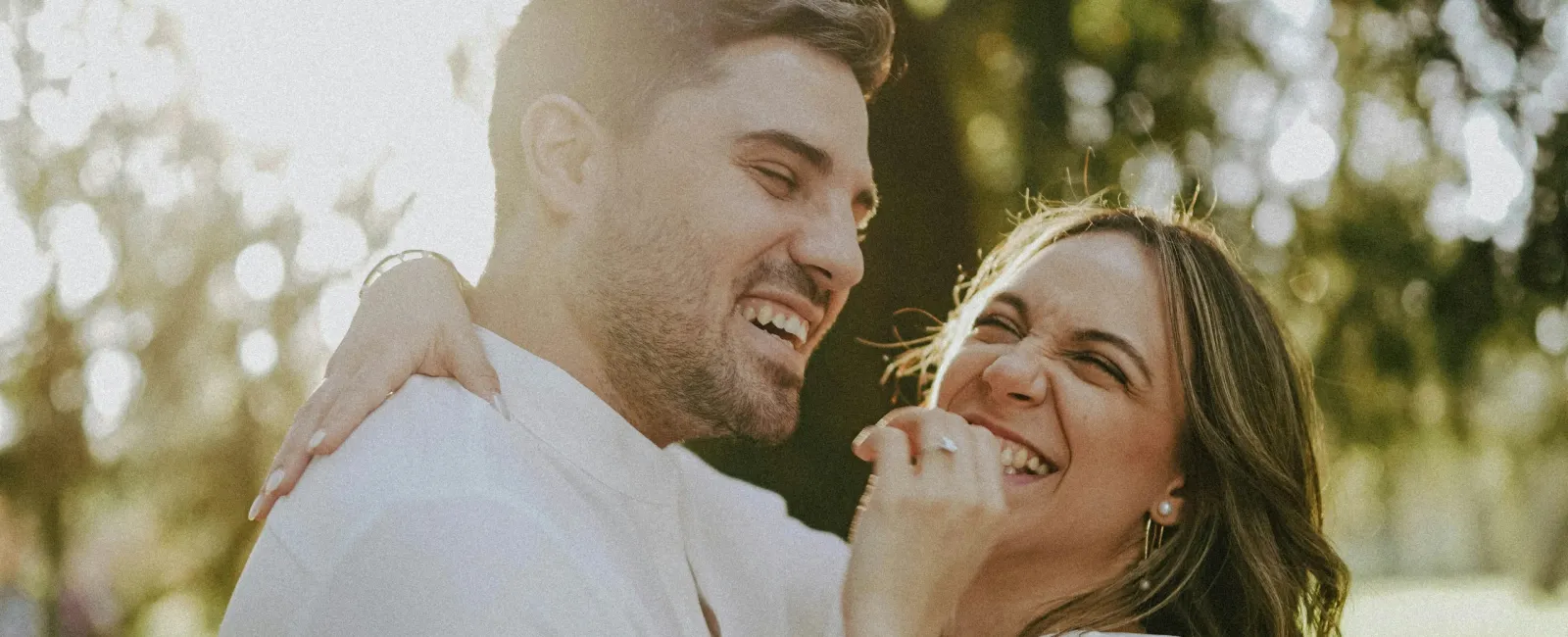 Happy couple embracing and laughing outdoors in bright natural light with green trees in the background