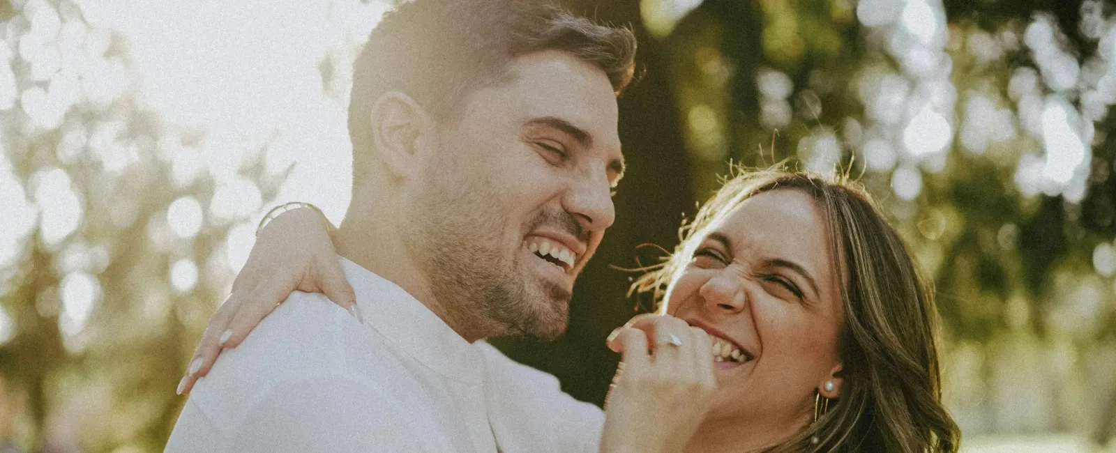 Happy couple embracing and laughing outdoors in bright natural light with green trees in the background