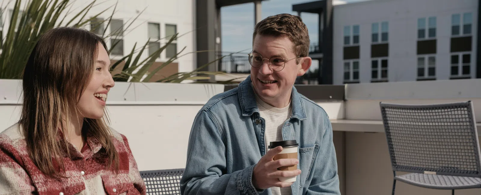 Two people chatting and smiling outdoors on a sunny day, one holding a coffee cup, casual urban setting.