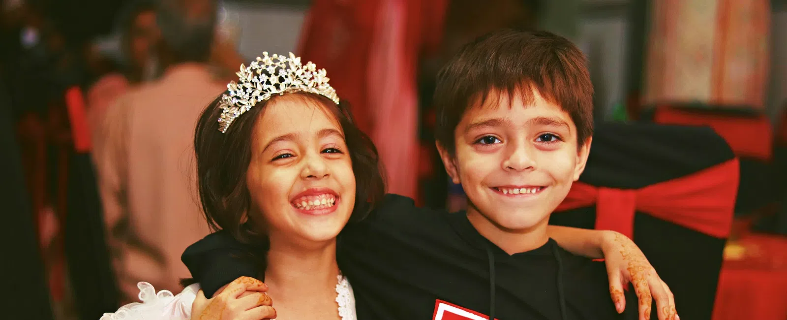 Smiling young girl in white dress and tiara hugs boy in black hoodie at festive indoor event.