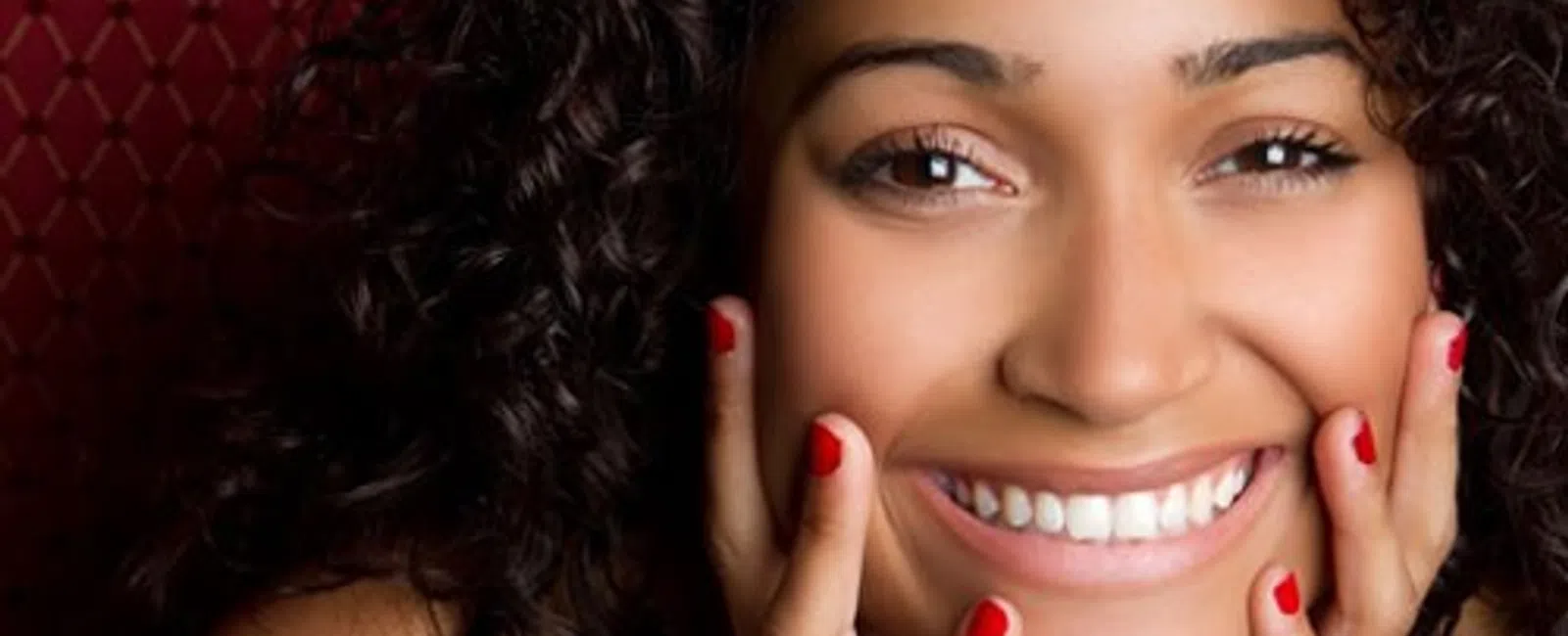Close-up of a smiling woman with curly hair and red nail polish touching her face against a red patterned background