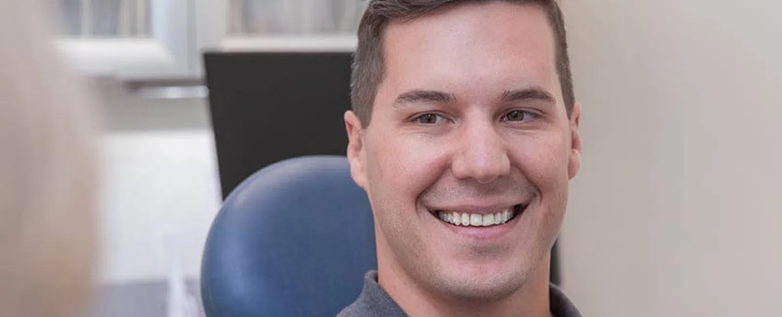 Smiling man sitting in a dental chair wearing a gray polo shirt in a bright dental office
