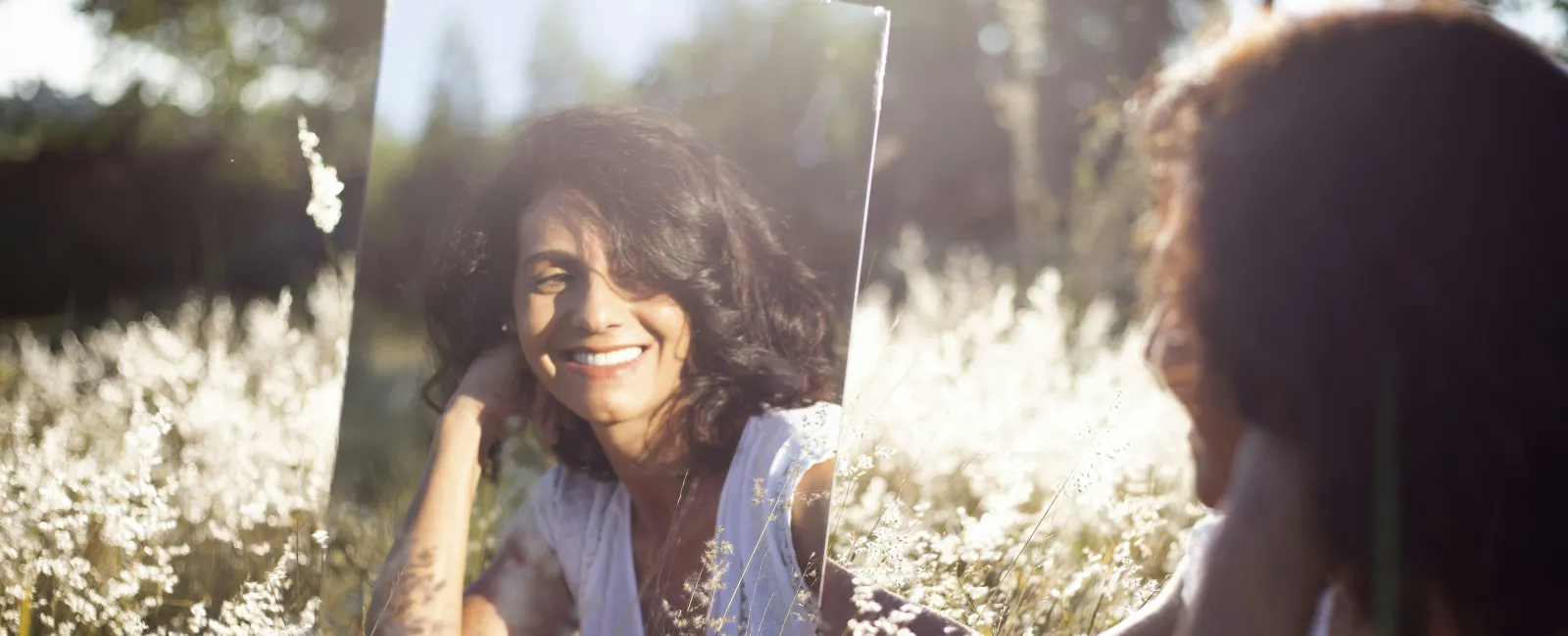 Woman smiling at her reflection in a mirror while sitting in a sunlit field of tall grass and wildflowers.