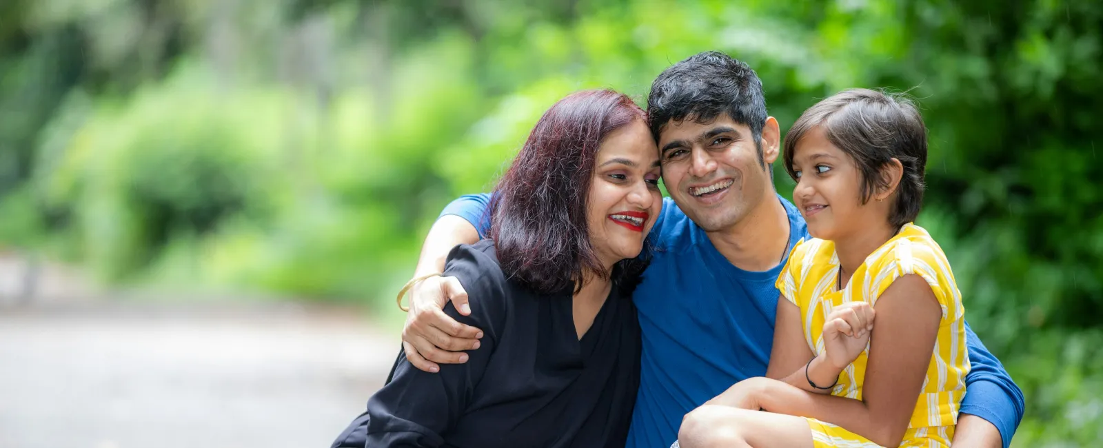 Smiling family of three sitting on a path outdoors surrounded by greenery, enjoying quality time together.