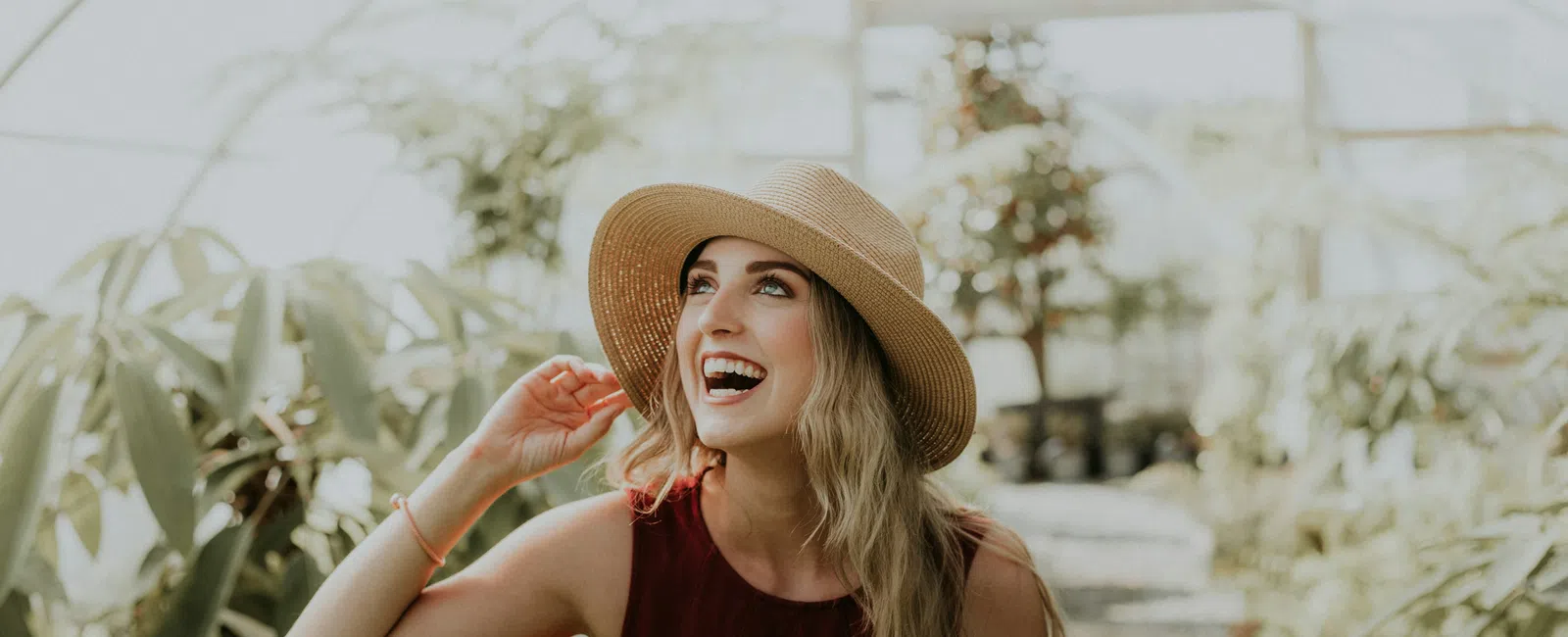Smiling young woman in straw hat and red dress enjoying a sunny day in a lush greenhouse.