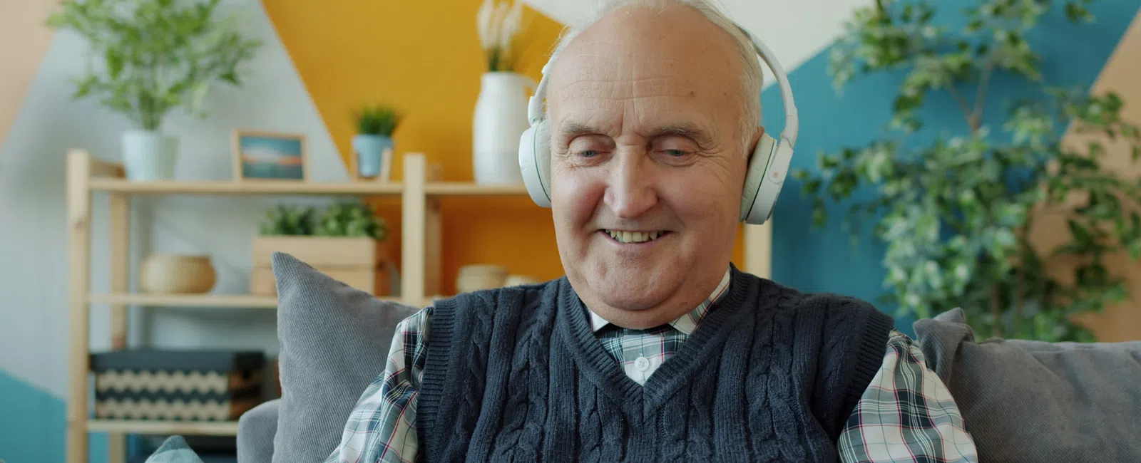 Senior man smiling while wearing headphones and using a tablet at home on a couch with colorful decor.