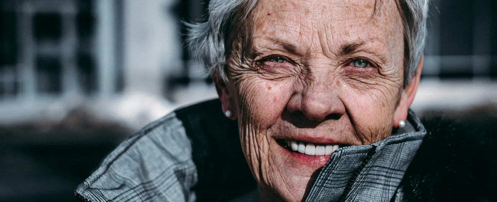 Smiling elderly woman with short gray hair wearing a gray jacket outdoors in natural light