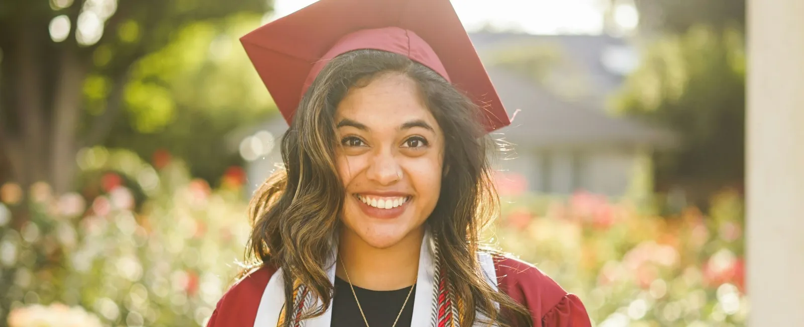 Smiling young woman in maroon graduation gown and cap with salutatorian stole outdoors in sunlight