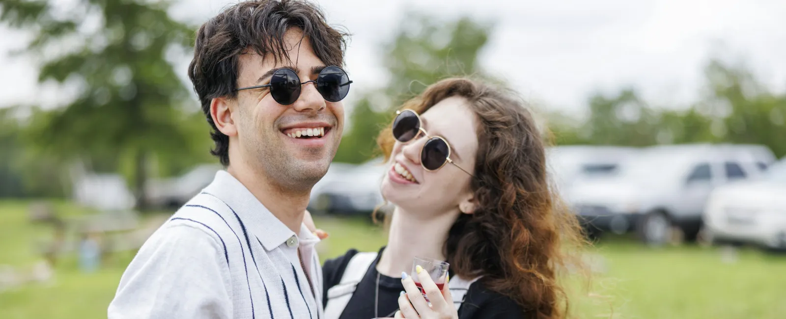 Smiling young couple in sunglasses enjoying outdoors holding drinks on a sunny day in a green park area.