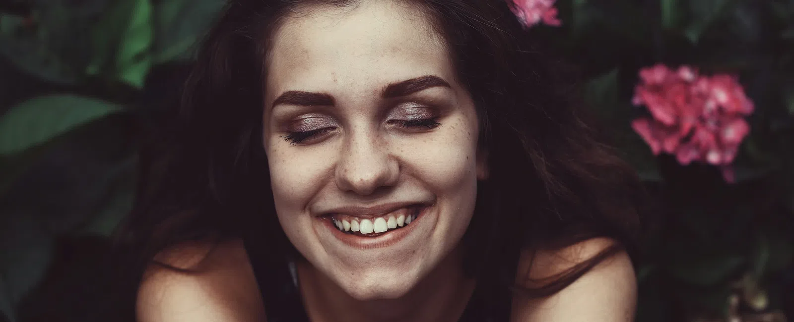 Young woman smiling with eyes closed surrounded by green leaves and pink flowers.