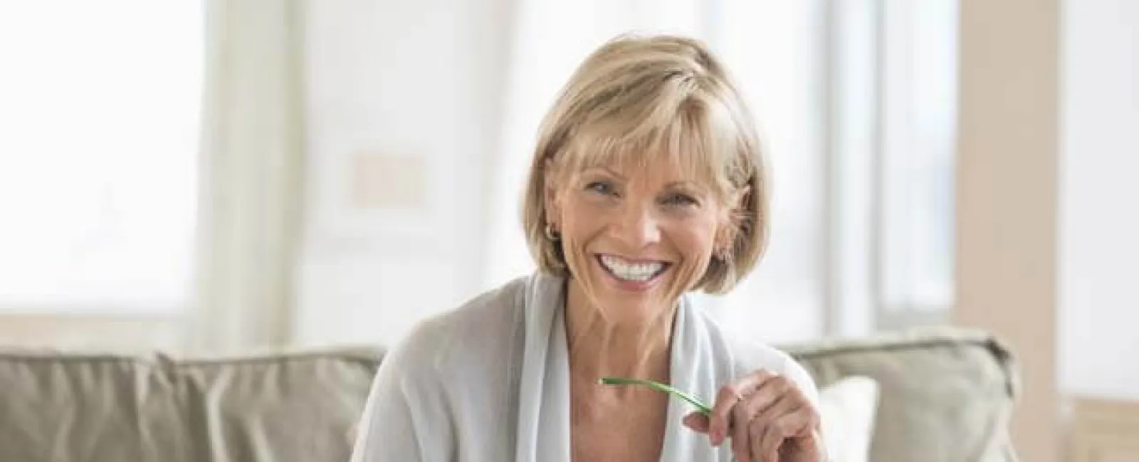 Smiling older woman using a laptop at home, sitting on a sofa with glasses in hand.