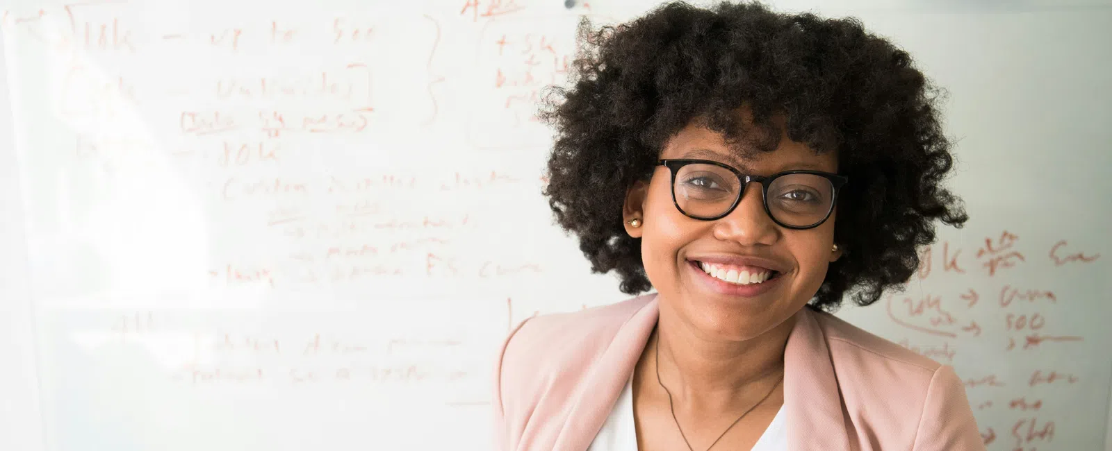 Smiling woman with glasses and curly hair in a pink blazer standing in front of a whiteboard with notes.