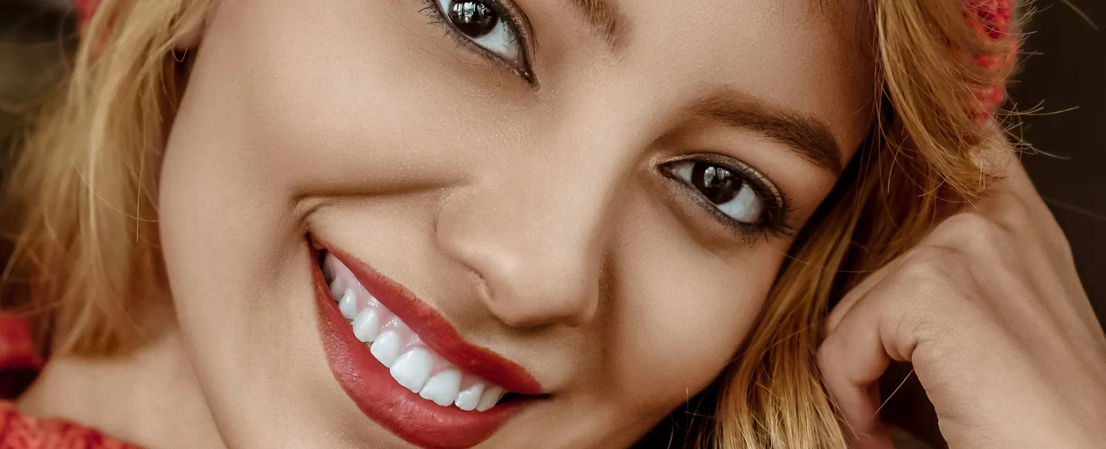 Smiling woman wearing a red knit hat and sweater with warm lights blurred in the background.