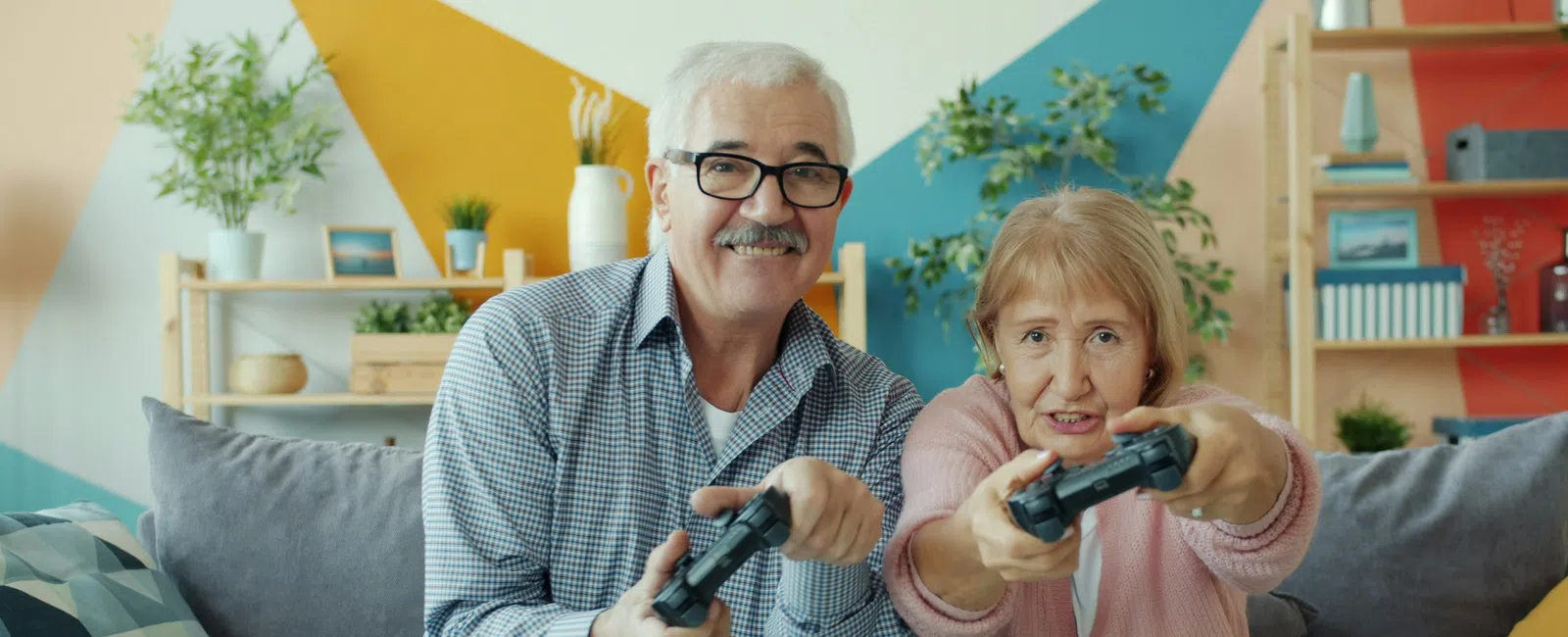 Senior couple smiling and playing video games together on a couch in a colorful living room.