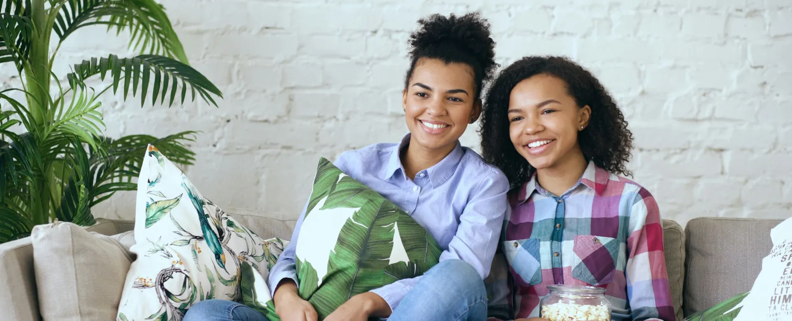 Two smiling young women sitting on a couch with tropical-themed pillows, enjoying popcorn and time together