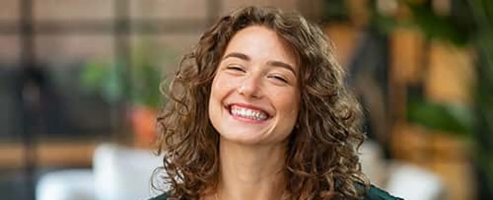 Smiling woman with curly hair wearing a dark green blouse and gold necklace in a bright indoor setting