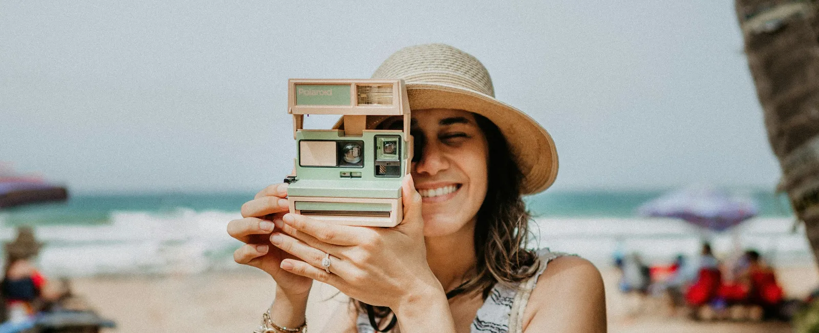 Smiling woman in striped top and hat holding vintage instant camera at beach with ocean in background