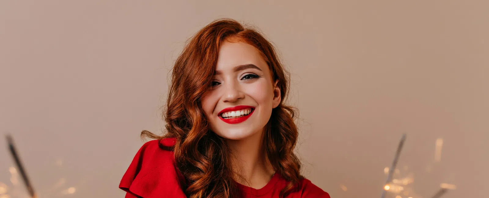 Smiling red-haired woman in a red dress holding sparklers celebrating with joy against a neutral background