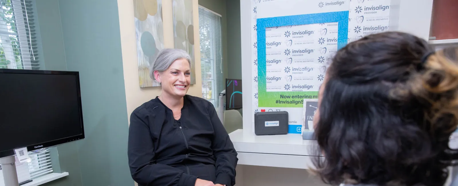 Smiling dental professional in black scrubs consulting a patient seated in a dental chair in a clinic room.