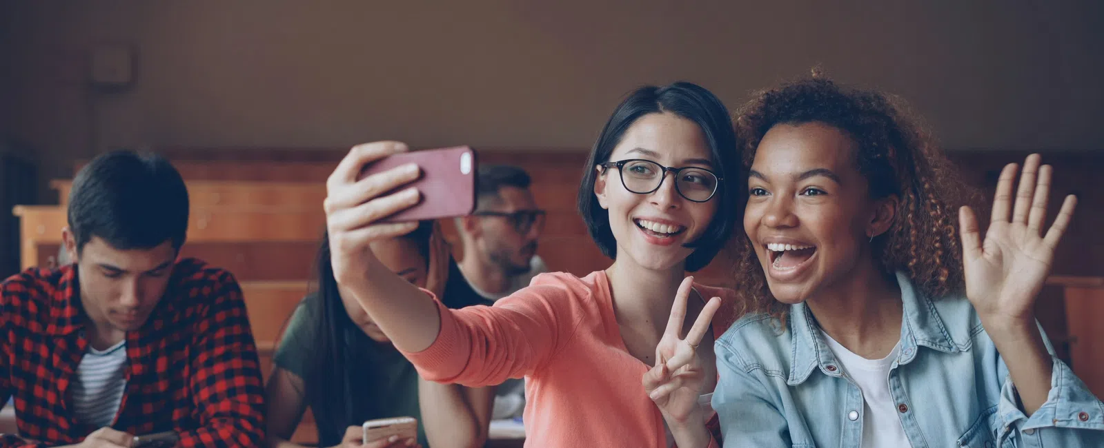 Two young women in a classroom taking a selfie, smiling and making hand gestures, with students and books in the background